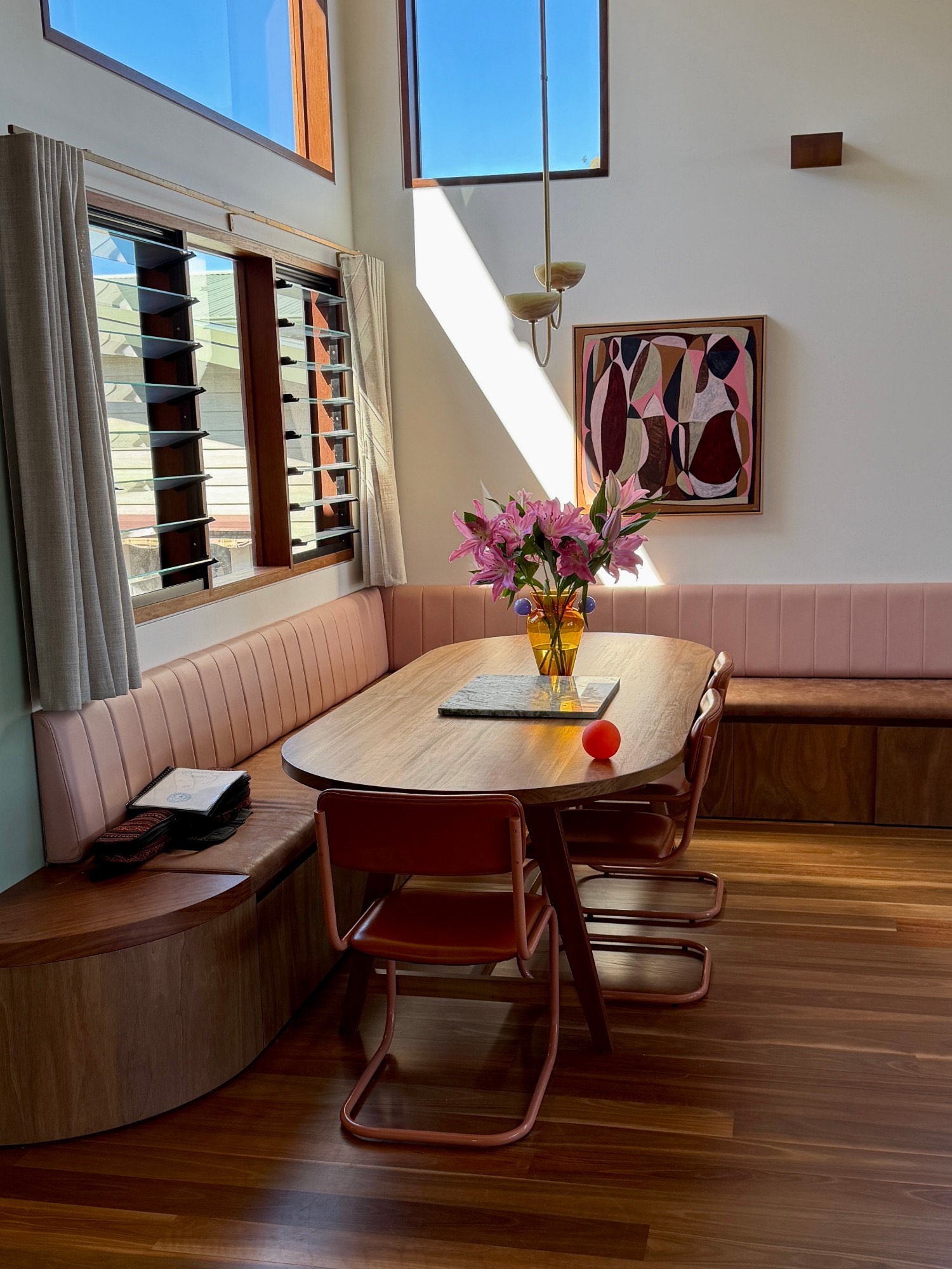 Sunlit dining nook with pink banquette seating around an oval timber table