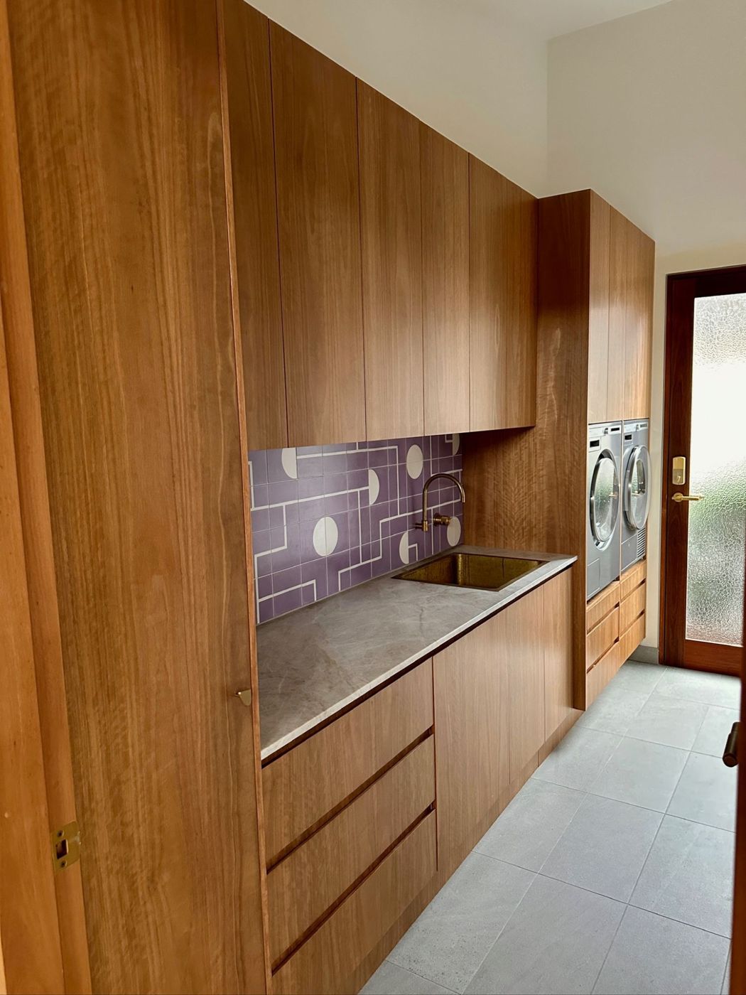 Timber laundry with full-height cabinetry, a stone benchtop, brass sink and raised washer and dryer